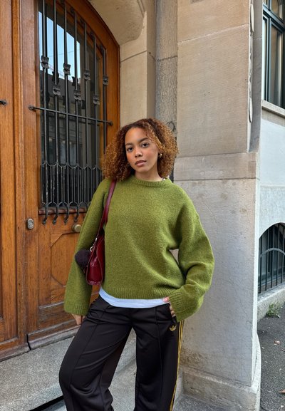 Suéter verde de gran tamaño hecho de tela texturizada, combinado con pantalones deportivos negros con rayas amarillas. Bolso rojo con detalle de pompón.