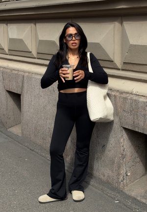 Mujer con ropa negra y gafas de sol sosteniendo café helado con pajita, de pie en la acera contra una pared de piedra texturizada, llevando una gran bolsa blanca.