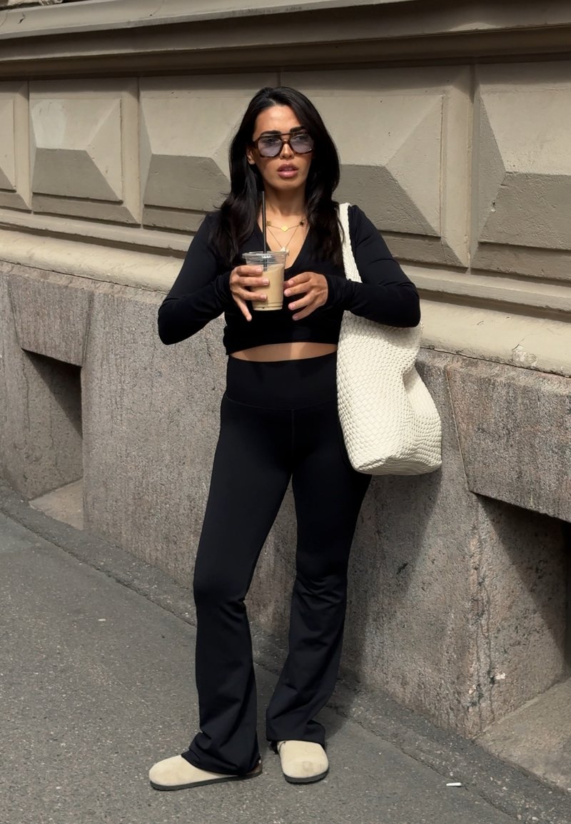 Woman in black outfit and sunglasses holding iced coffee with straw, standing on sidewalk against textured stone wall, carrying large white bag.