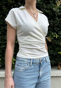 Λευκή μπλούζα με κοντά μανίκια και λεπτομέρεια στο γιακά, από απαλό βαμβακερό ύφασμα, συνδυασμένη με ψηλόμεσα ανοιχτό μπλε τζιν και ασημένιο κολιέ.