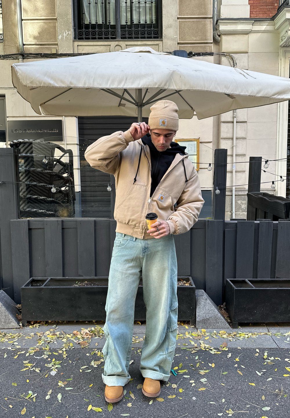 Beige hoodie met een voorzak, zwarte binnenlaag, lichtblauwe baggy jeans, tan kleurige laarzen, met een koffie kop in de hand, onder een parasol.