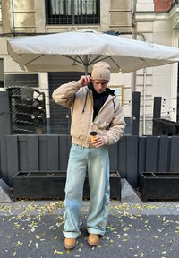 Beige hoodie met een voorzak, zwarte binnenlaag, lichtblauwe baggy jeans, tan kleurige laarzen, met een koffie kop in de hand, onder een parasol.