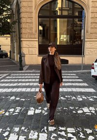 Hnedý dokonale strihaný blejzer na zodpovedajúcom oblečení, hnedá taška a baseballová čiapka. Kráčajúci po kamennej ulici s bielymi priečkami.