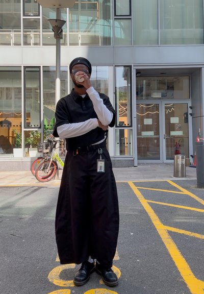 Camisa polo negra de manga corta sobre una camisa blanca de manga larga, combinada con pantalones largos negros. Lleva gafas y sostiene una bebida.