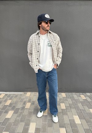 Hombre con gafas de sol, gorra, camisa de cuadros sobre camiseta gráfica, vaqueros azules y zapatillas blancas, de pie sobre pavimento de baldosas frente a una pared gris.