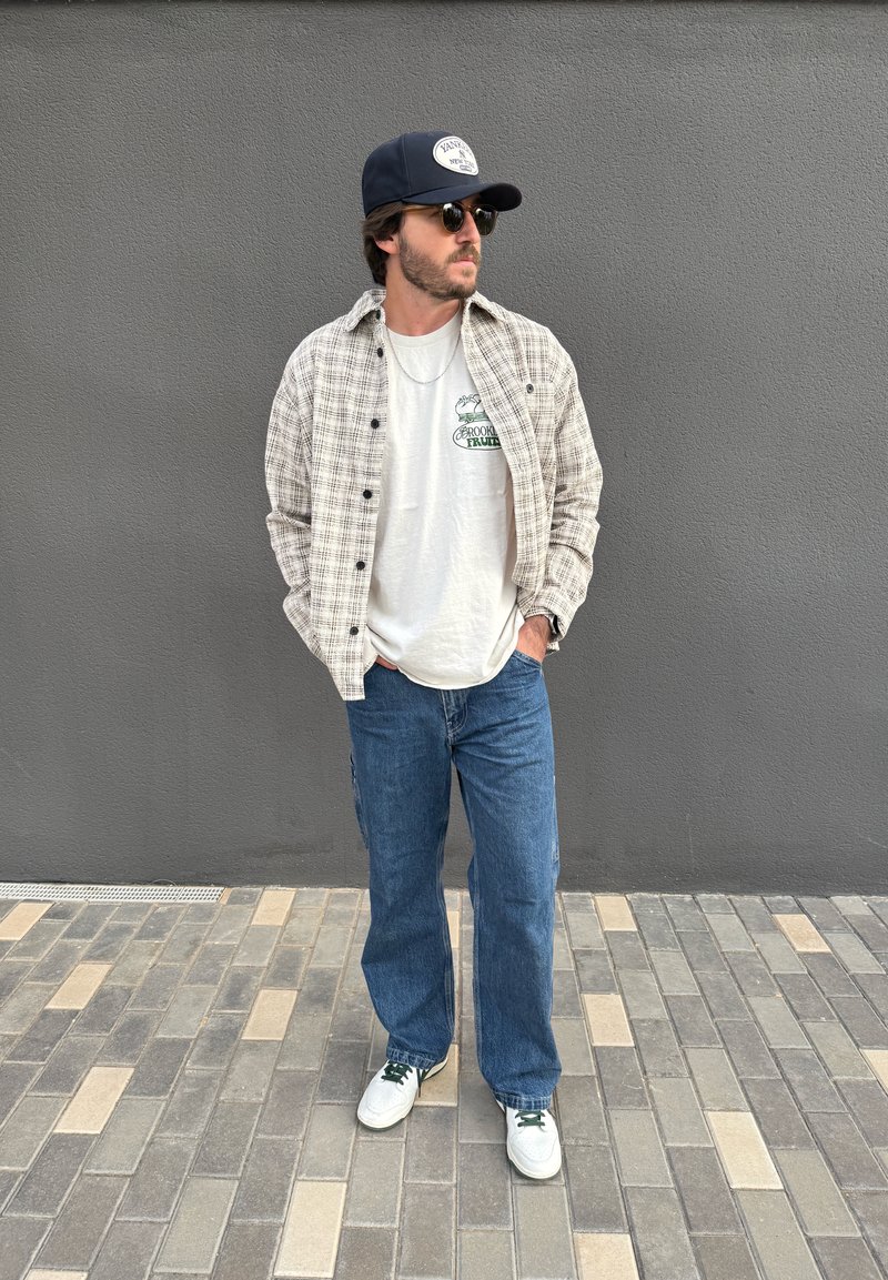Man wearing sunglasses, a cap, a checkered shirt over a graphic t-shirt, blue jeans, and white trainers standing on tiled pavement against a grey wall.