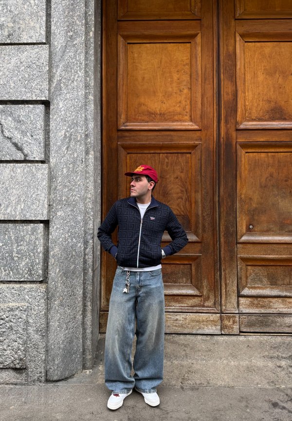 Man i röd keps, svart mönstrad jacka, vida jeans och vita sneakers står med händerna i fickorna mot en stor träport.