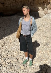 Νέος άνδρας με γκρι αμάνικο μπλουζάκι, ανοιχτό ριγέ πουκάμισο, μαύρα σορτς, πράσινα αθλητικά παπούτσια και ταμπά τσάντα ώμου στέκεται σε βραχώδες έδαφος κάτω από τον ήλιο κοντά στην είσοδο σπηλιάς.