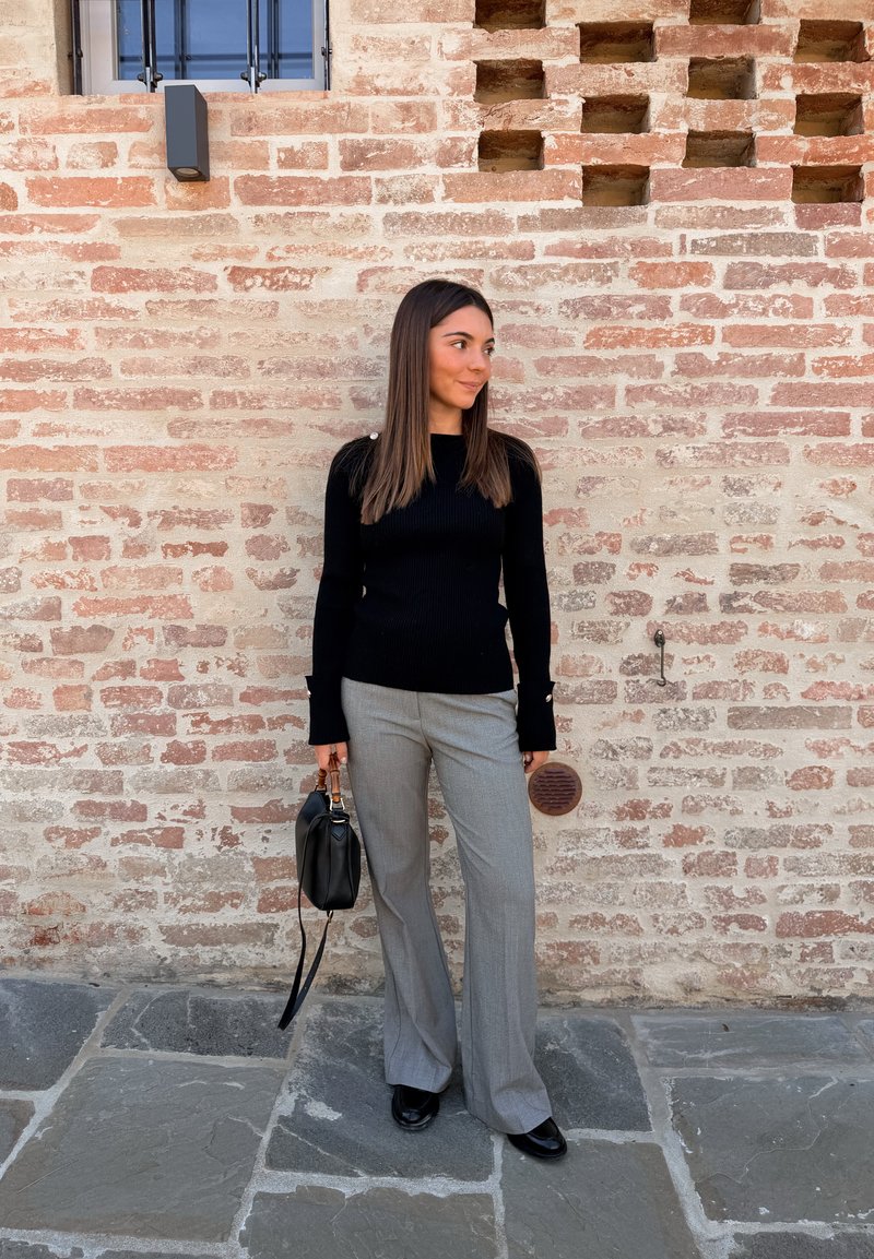 Schwarzer gerippter Pullover mit langen Ärmeln, graue weit geschnittene Hose und eine kleine schwarze Handtasche, vor einer strukturierten Ziegelwand.