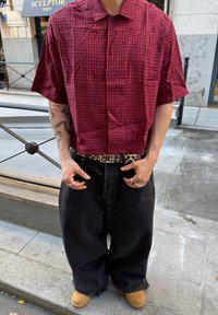 Camicia a maniche corte a scacchi rossi e neri con colletto abbottonato, abbinata a pantaloni in denim neri larghi e una cintura con stampa leopardata.