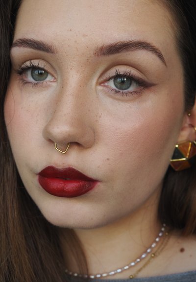Primer plano de una cara con piel marrón clara, ojos verdes y labios carnosos que llevan lápiz labial rojo oscuro. Anillo de septum dorado y pendientes geométricos.