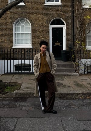 Joven de pie en la calle frente a una casa de ladrillo con puerta negra arqueada, vistiendo un abrigo beige, suéter mostaza y pantalones oscuros con rayas blancas.