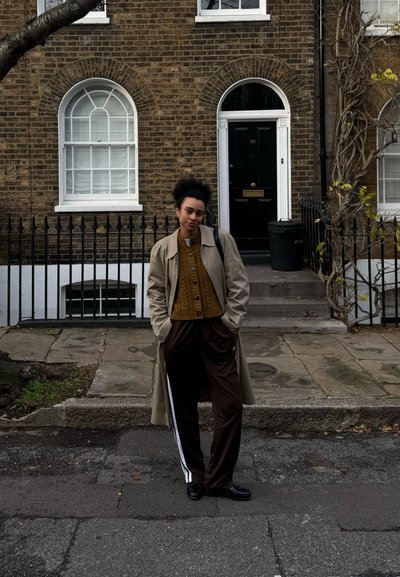 Joven de pie en la calle frente a una casa de ladrillo con puerta negra arqueada, vistiendo un abrigo beige, suéter mostaza y pantalones oscuros con rayas blancas.