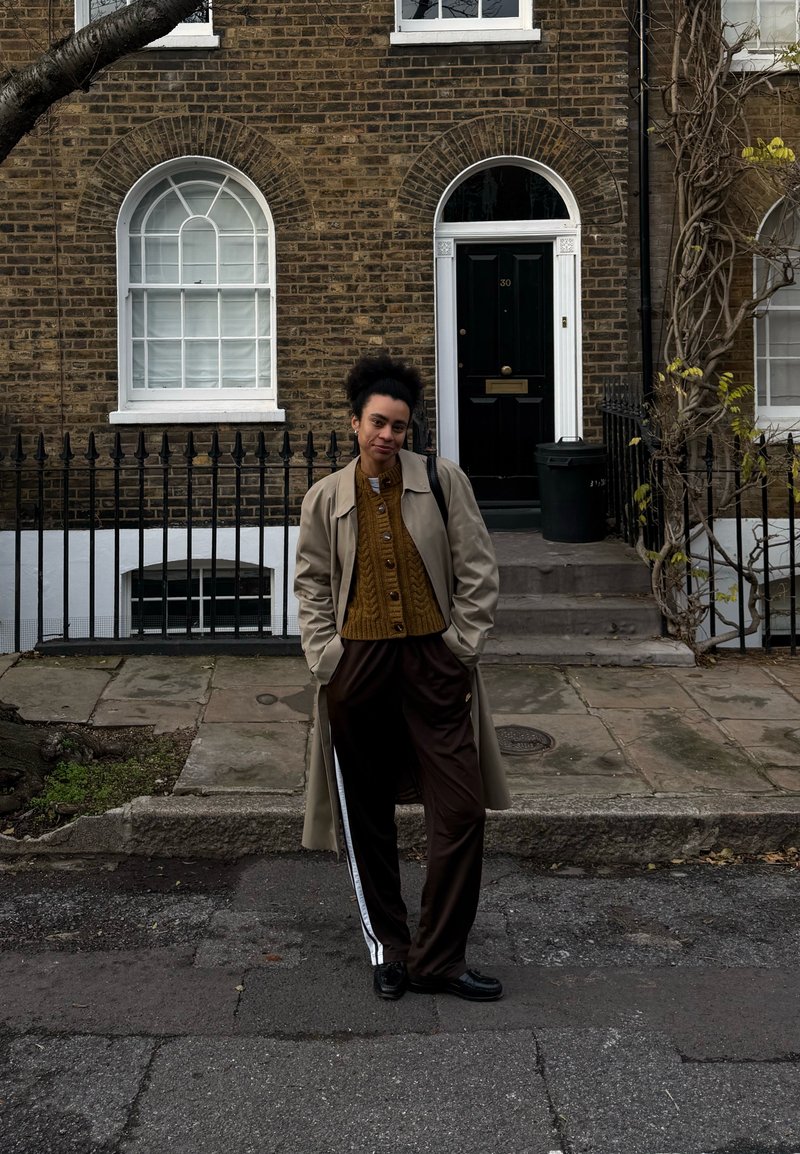 Jeune personne debout dans la rue devant une maison en briques avec une porte noire arquée, portant un manteau beige, un pull moutarde et un pantalon foncé avec des rayures blanches.