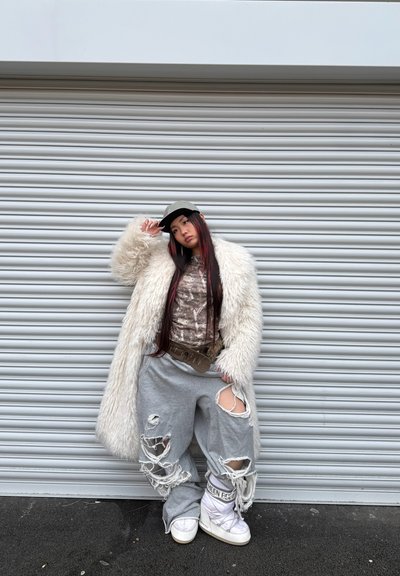 Abrigo de piel sintética blanca, pantalones de chándal grises rasgados, blusa estampada. Zapatillas deportivas blancas de caña alta. Gorra gris, cabello largo oscuro con reflejos rojos.