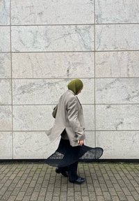 Kvinne i grønn hijab, grå lang blazer og svart kjole, går på steinlagt fortau ved siden av en teksturert lys grå vegg. Lys og skygge er til stede.