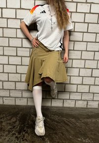 Weisse Adidas-Jersey mit roten und schwarzen Akzenten, khakifarbenen Rock mit Knopfdetails, weissen kniehohen Socken und grauen Turnschuhen. Backstein-Hintergrund.