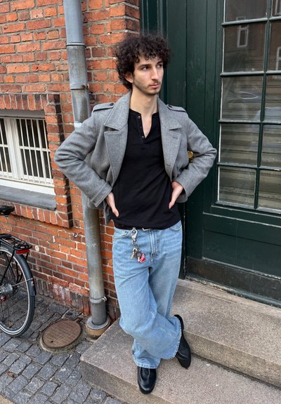 Chaqueta gris corta de doble botonadura, camiseta de manga larga negra, jeans de pierna ancha azul claro y zapatos negros. Un llavero cuelga de la cinturilla.