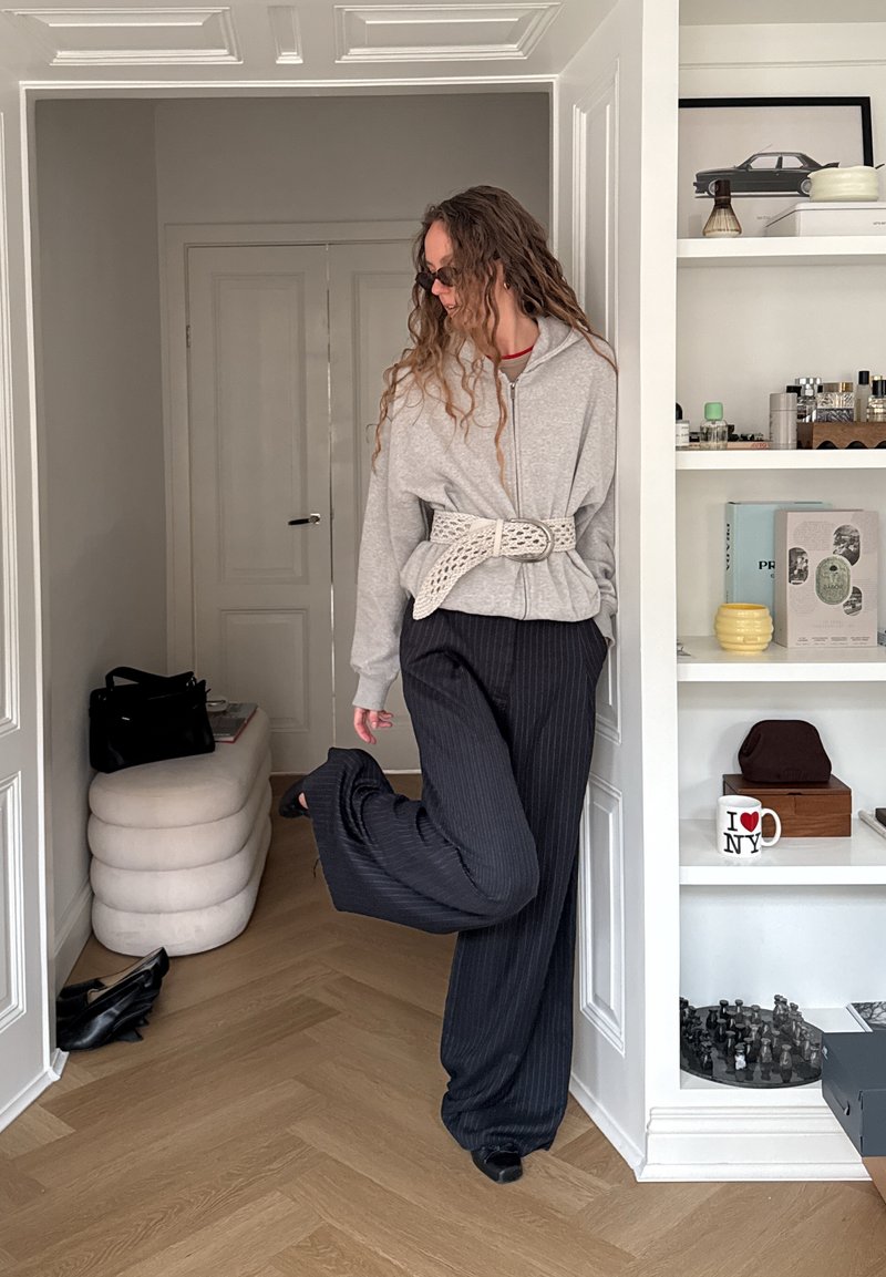 Femme en sweat à capuche gris avec une large ceinture et un pantalon à fines rayures, posant avec une jambe levée dans une pièce lumineuse avec des étagères et un petit pouf.