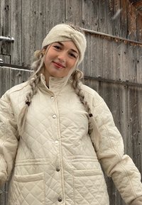 Giacca imbottita crema con chiusura a bottone a scatto e due tasche frontali. Il modello indossa un cerchietto beige; fiocchi di neve visibili sullo sfondo.