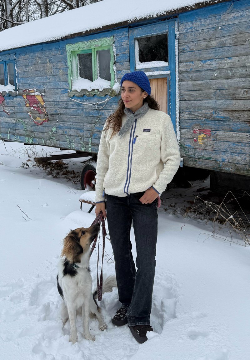 Veste en polaire crème avec des accents bleus et une poche avant, associée à un jean foncé. Un chien brun et blanc est assis à côté de la personne dans la neige.