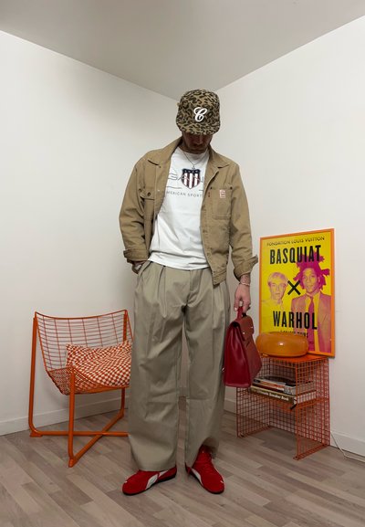 Hombre con gorra de leopardo, chaqueta beige, camisa blanca, pantalones anchos beige, zapatos rojos, sosteniendo un bolso rojo en una habitación con silla naranja y póster de Basquiat Warhol.