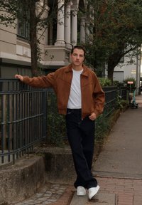 Giacca in suede marrone con colletto, abbinata a jeans blu scuro e sneakers bianche, in piedi contro un parapetto in un contesto urbano.