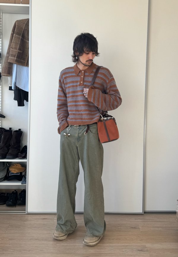 Homme portant un pull rayé marron et gris, un pantalon large vert, des chaussures beige, et tenant un sac à main orange, debout dans une pièce avec des étagères.