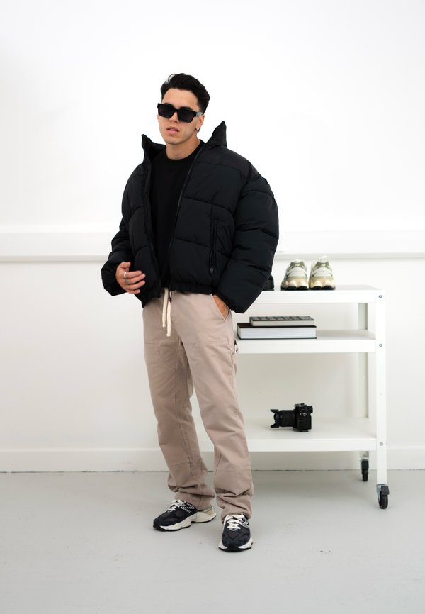 Nero giubbotto puffo con un design cropped, pantaloni cargo beige e sneaker nere. Gli accessori includono occhiali da sole e anelli in argento.