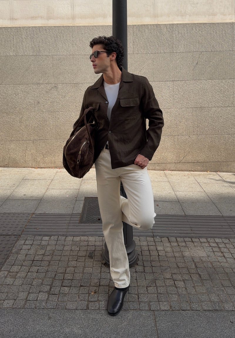 Jeune homme en lunettes de soleil appuyé contre un réverbère, portant une veste foncée, une chemise blanche, un pantalon crème, des chaussures noires et tenant un sac marron.