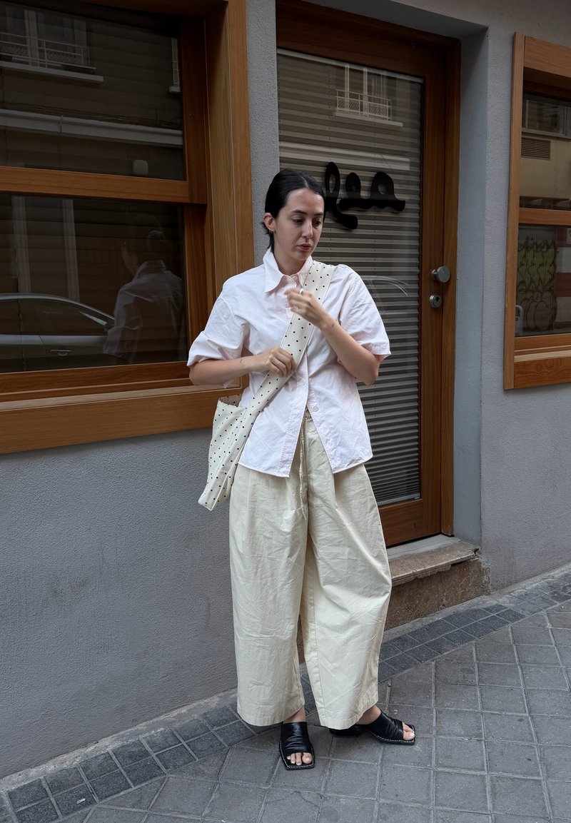 Femme debout sur le trottoir ajustant la bride d'un sac fourre-tout blanc à pois, portant une chemise blanche à manches courtes, un pantalon large crème et des sandales noires.