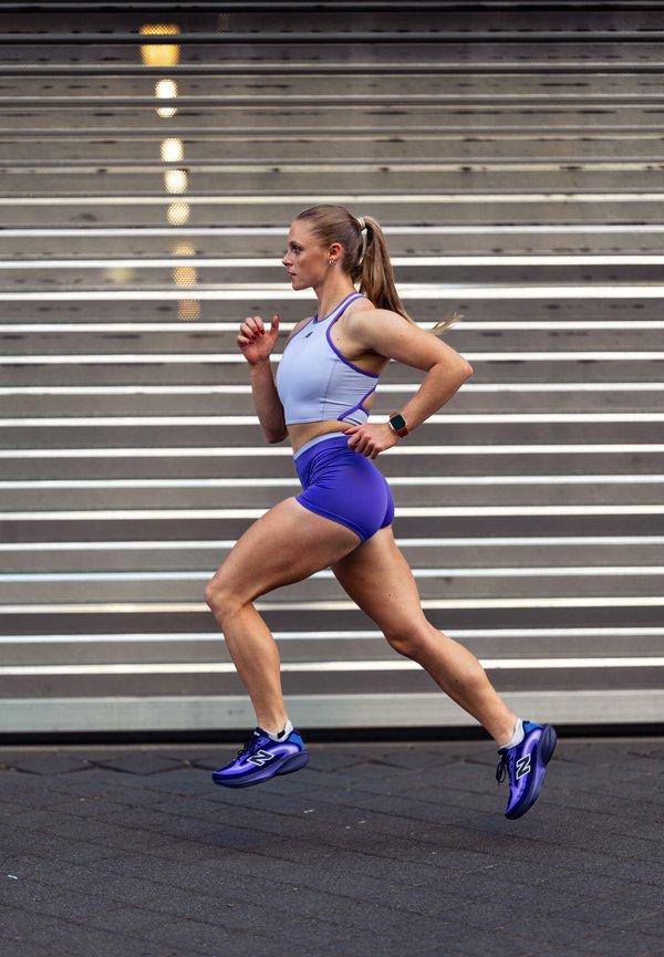 Femme athlétique en tenue de course bleue sprintant sur le trottoir devant un rideau métallique, en pleine foulée, regard concentré vers l'avant.