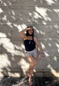 Top negro de un solo hombro, shorts a rayas blancas y chanclas marrones; de pie frente a una pared de mármol claro con patrones de sombra.