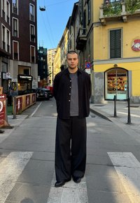 Crna jakna preko sive majice, široke crne hlače, stoji na gradskoj ulici. Okolne zgrade su od različitih materijala i boja, sa uličnim znakovima.