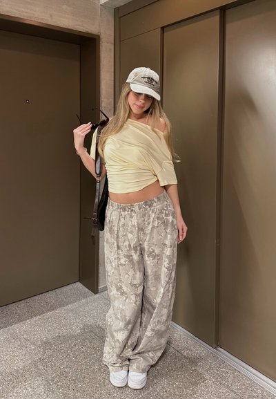 Joven mujer con pantalones sueltos de estampado, top crema sin hombros, gorra blanca, zapatillas blancas, sosteniendo gafas de sol y bolso negro en pasillo con puertas cerradas.