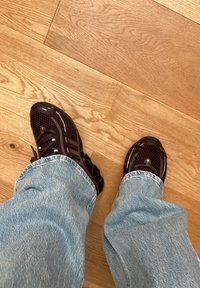 Baskets de sport marron foncé avec du matériau en mesh, accents réfléchissants et un système de laçage, associées à un jean bleu clair sur un sol en bois.