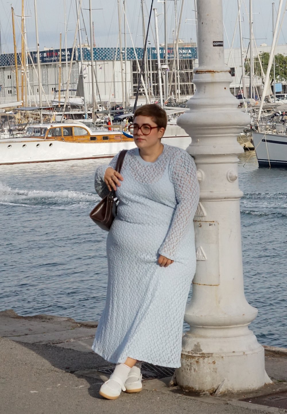 Vestido longo azul claro, texturizado, com mangas longas, combinado com meias brancas e sapatos claros, posando junto a um candeeiro branco perto da água.