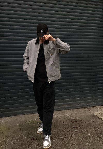 Chaqueta gris con cuello negro, llevada sobre una camiseta negra. Pantalones negros y zapatillas grises con acentos blancos. Gorro negro con logotipo.