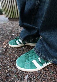Baskets en daim vert avec des rayures blanches, dotées d'un bout rond et d'une semelle plate en caoutchouc, portées avec un jean en denim foncé. La texture du sol est visible.