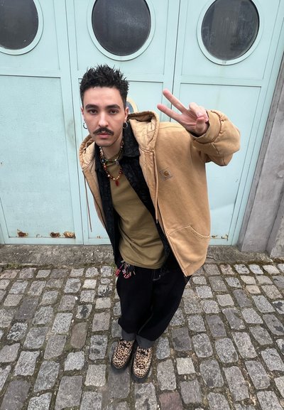 Chaqueta con capucha marrón, camiseta negra, blusa verde oliva, pantalones negros y zapatos de estampado de leopardo. Collares de cuentas y accesorios visibles. Fondo de ladrillo.