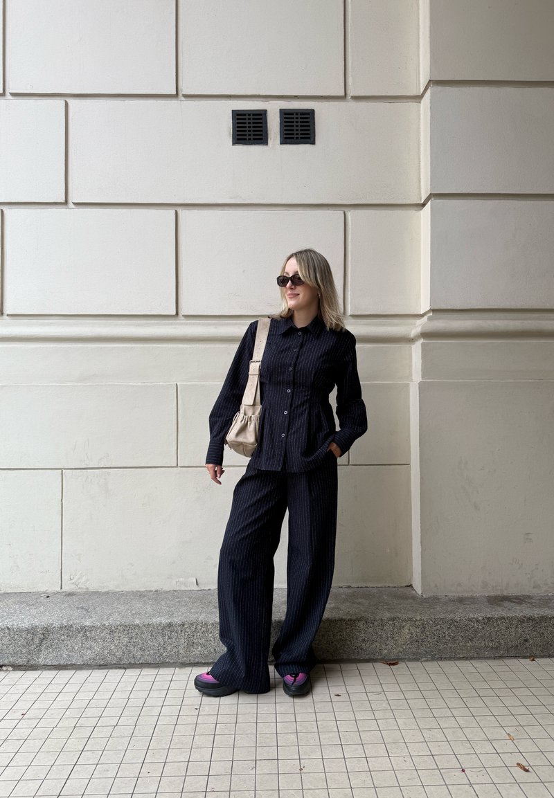 Chemise boutonnée noire à rayures avec un pantalon large assorti, accompagnée d'un sac beige clair et de chaussures noires accentuées de rose, devant un mur blanc.