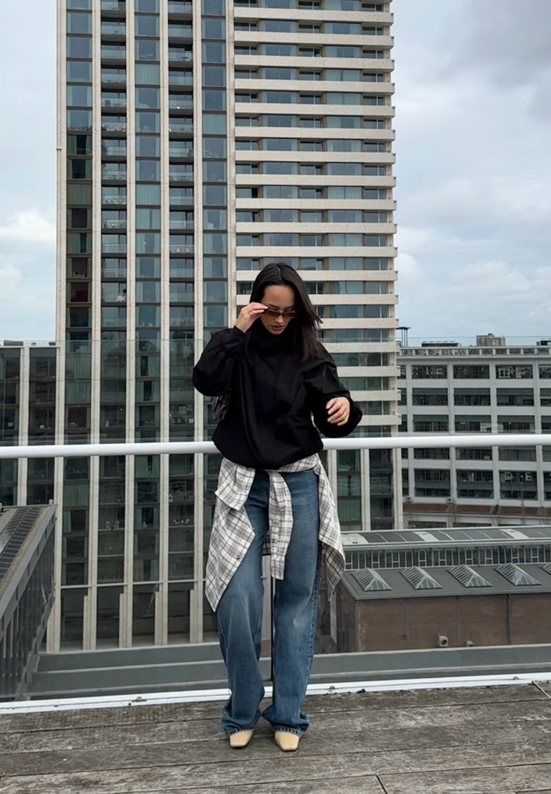 Felpa nera oversize con maniche lunghe, abbinata a jeans azzurri e a una camicia a quadri annodata in vita. Scarpe beige a punta.