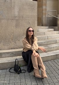 Beżowy, oversize sweter, czarna, zwiewna spódnica i brązowe kozaki do kolan. Mała czarna torebka na kamiennych schodach o teksturowanej powierzchni.