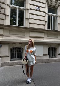 Camisa branca de mangas curtas com estampas de conchas laranjas, combinada com calções brancos de renda. Mala preta e bege, sapatos de bolinhas e meias brancas.