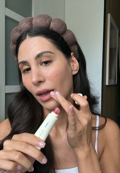 Bálsamo labial en un tubo suave y redondeado con un aplicador rosa-rojo. La persona lo usa para aplicar color a sus labios, llevando una diadema marrón y texturizada.