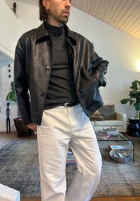 Czarna skórzana kurtka z kołnierzem i guzikami, noszona na czarnym golfie, w połączeniu z białymi spodniami. Wnętrze z roślinami i meblami.