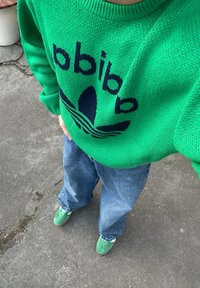 Πράσινο πλεκτό πουλόβερ με ναυτικό λογότυπο, χαλαρές μπλε τζην παντελόνες και πράσινα σουέτ παπούτσια. Υφή υφάσματος και άνετη εφαρμογή. Η επιφάνεια του εδάφους είναι γκρι.