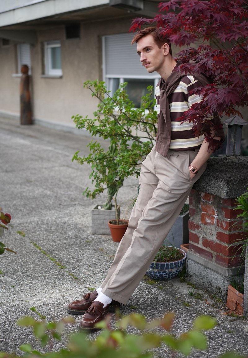 Jeune homme en chemise rayée, pantalon beige et chaussures marron, appuyé contre un muret en briques près de plantes en pot et d'un arbuste à feuilles rouges en extérieur.