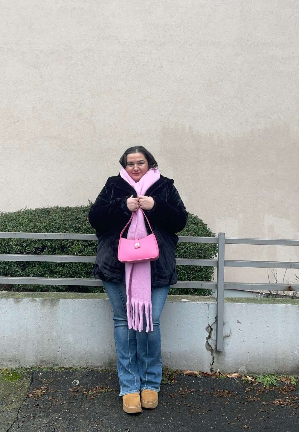 Sort pelstrøje, lyserød tørklæde med frynser, lille lyserød håndtaske, blå jeans, beige støvler; står mod en simpel væg med grønne planter.