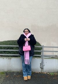 Sort pelstrøje, lyserød tørklæde med frynser, lille lyserød håndtaske, blå jeans, beige støvler; står mod en simpel væg med grønne planter.